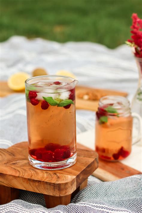 Un pichet de thé glacé garni de framboises fraîches, de feuilles de menthe et de tranches de citron sur une table en bois ensoleillée.